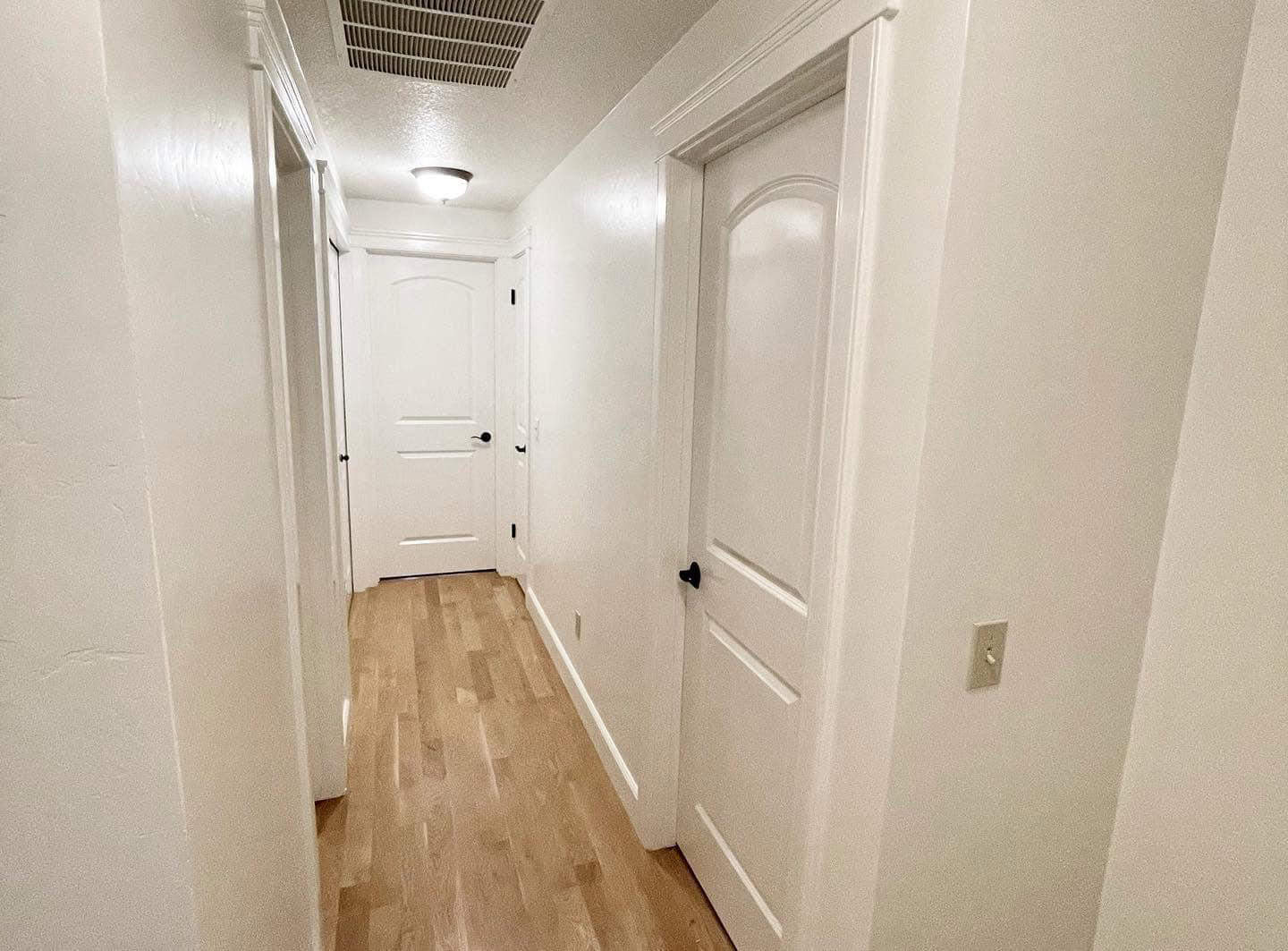 A bright and airy hallway with white walls and a wooden floor. There are several doors leading to different rooms, and a light fixture hangs from the ceiling.