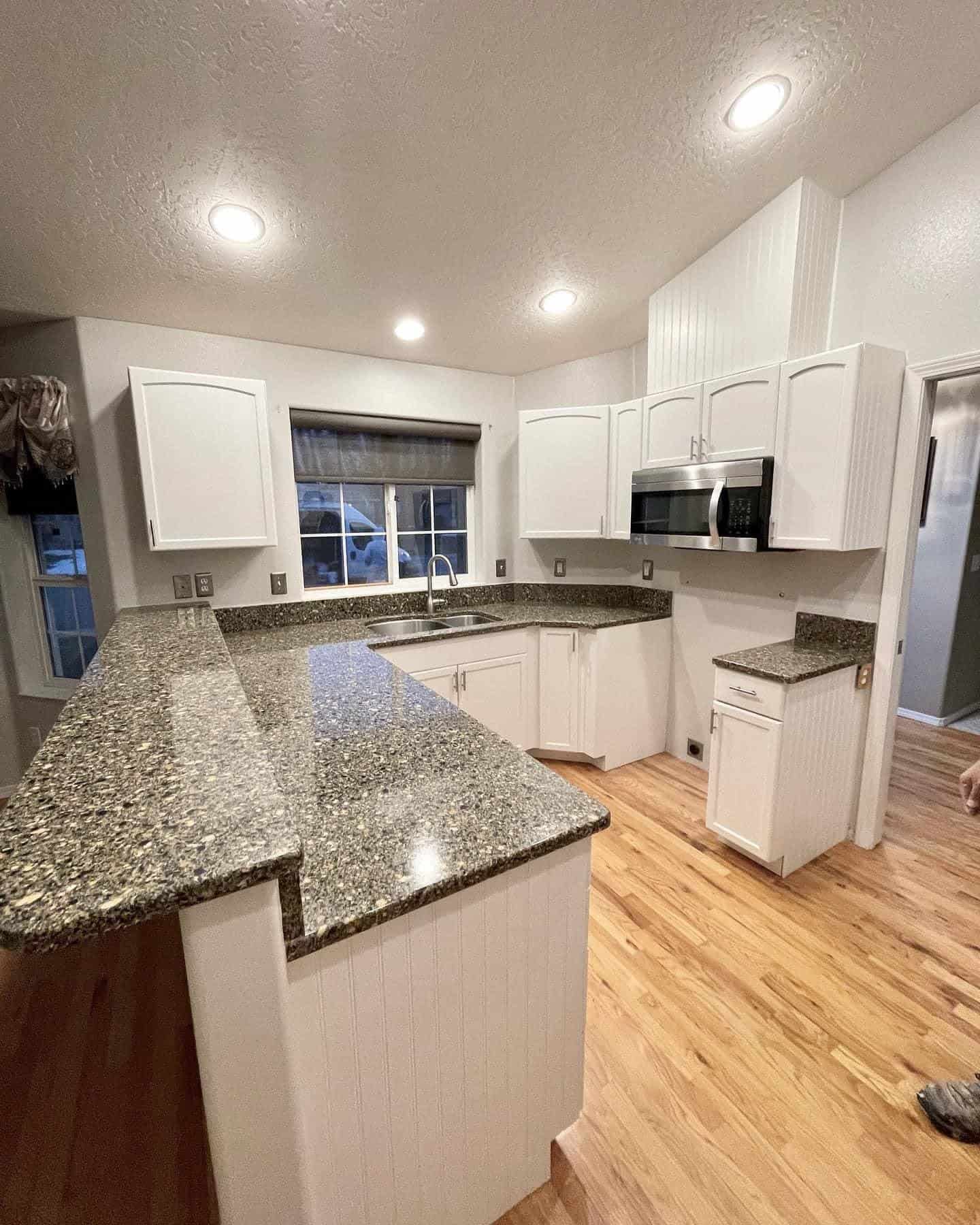 A kitchen with white cabinets, a large granite countertop island, and a wood floor. The kitchen features a microwave above the stove, a large window, and recessed lighting.